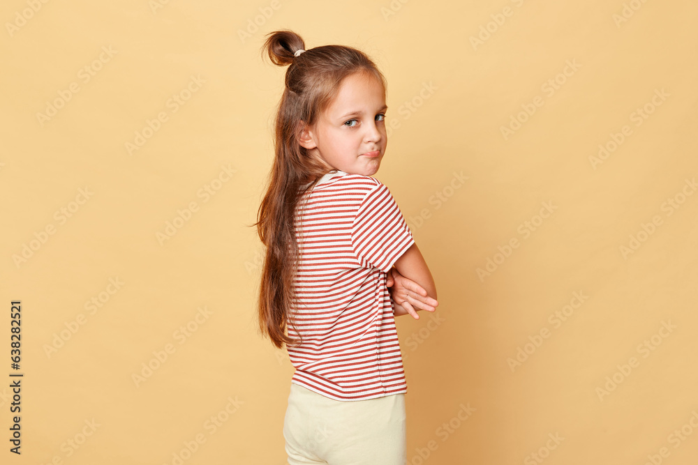 Offended sad dark haired little girl wearing striped T-shirt isolated over beige background turning away crossed hands looking at camera expressing negative feelings.