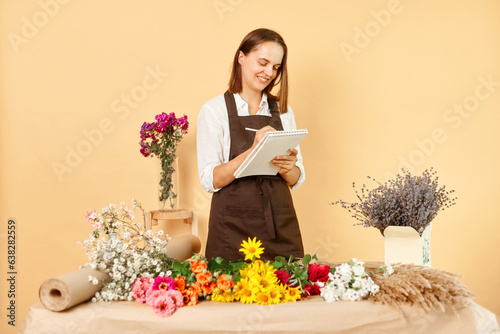 Cheerful woman wearing brown apron making notes receiving order in florist shop with various flowers smiling happily standing isolated over beige background.