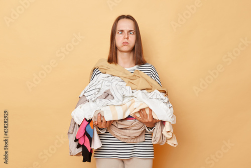 Tired exhausted brunette woman holding pile of clothes in hands isolated over beige background looking at camera with crossed eyes blowing cheeks doing lots house work. photo