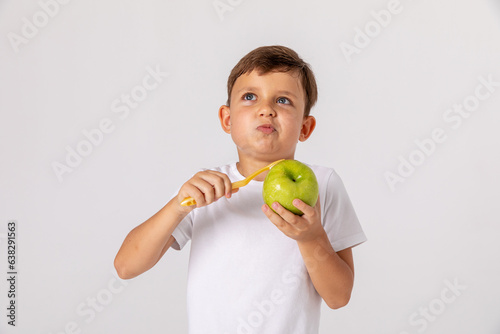 Cute thoughtful little boy in white t-shirt peeling a green apple with a toothbrush on white studio background. Dental care and health. Pediatric dentistry concept. Mock up. Space for text.