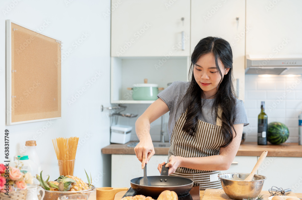 A beautiful young Asian girl in an apron is making pancakes in a minimalist home kitchen.