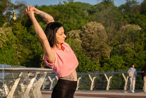 Lady raises both arms above head while warning up on bridge at sunrise. Sports activity in crowded place with bike path on warm day closeup © SlavaStock