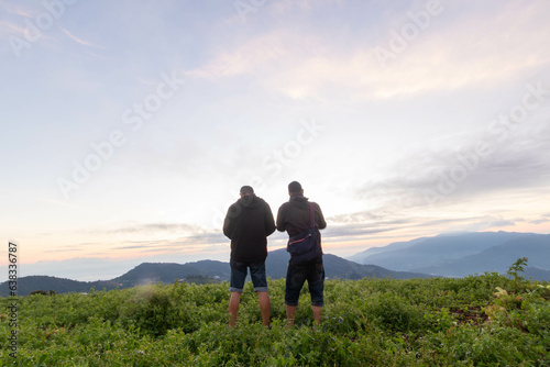 Male tourists stand on top of a mountain in the morning looking at the beautiful view of the twilight as the sun rises on the horizon and praying to God for blessings according to the Christian faith.