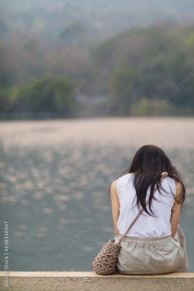 young woman sits on walkway by reservoir watching beautiful scenery of reservoir alone in the evening. back of woman sitting by reservoir alone in evening and with copy space for text. young woman sits on walkway by reservoir watching beautiful scenery of reservoir alone in the evening. back of woman sitting by reservoir alone in evening and with copy space for text.