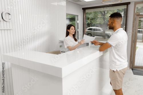 At reception desk beautiful medical assistant giving some blank information to the patient to signed before go to the doctor