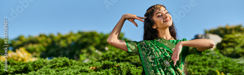 cheerful indian woman in matha patti and sari posing near plants with blue sky on background, banner