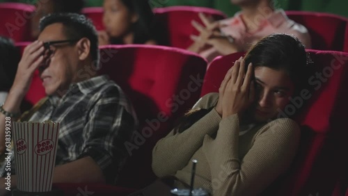 Asian man and woman having shocked and frightened reaction while watching horror thriller movie in the movie theater. Scared woman and man covering eyes when watching horror movie