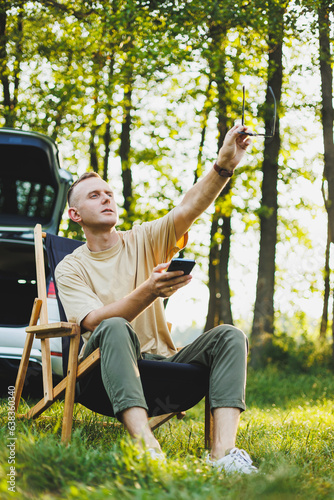 Freelancer man sits on a wooden chair in nature and works online on a laptop. A man travels and works remotely on a laptop computer. Office work in nature. Vacation