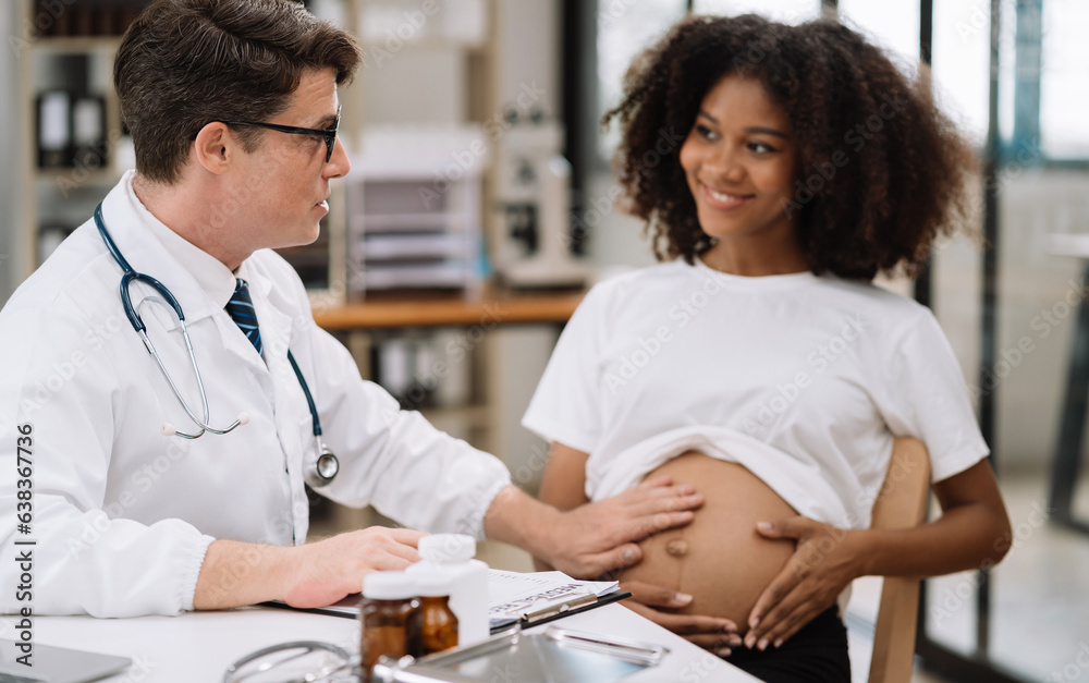 Obraz premium Happy pregnant woman has appointment with doctor at clinic. male medic specialist with stethoscope listens to baby's heartbeat in mother's belly. Pregnancy, health care concept.