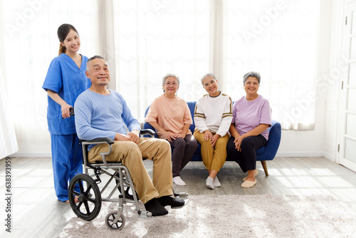 The caregiver therapist stands with an Asian senior sitting in a wheelchair with a group of senior women sitting on a sofa in the background. The nursing home facilitates a support group.