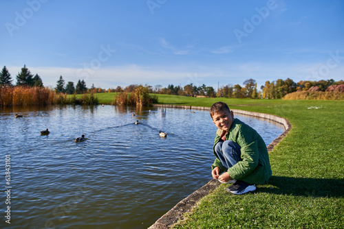 happy african american boy in outerwear and jeans sitting near pond with ducks, nature and kid © LIGHTFIELD STUDIOS