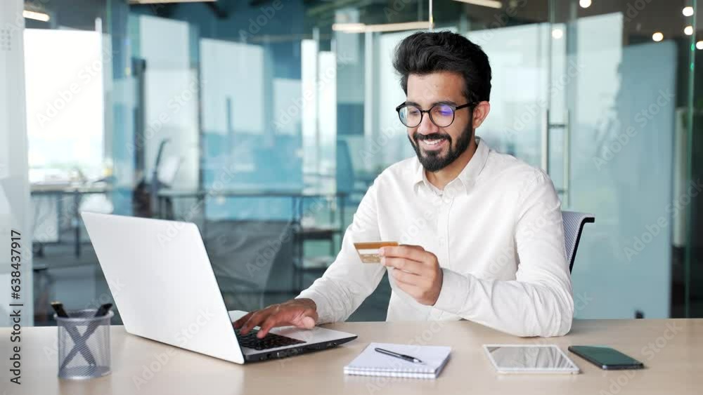 A smiling young bearded man enters a credit card number on a laptop while sitting at a desk at a workplace in a modern office. Handsome businessman in glasses shopping online, happy about good price A smiling young bearded man enters a credit card number on a laptop while sitting at a desk at a workplace in a modern office. Handsome businessman in glasses shopping online, happy about good price