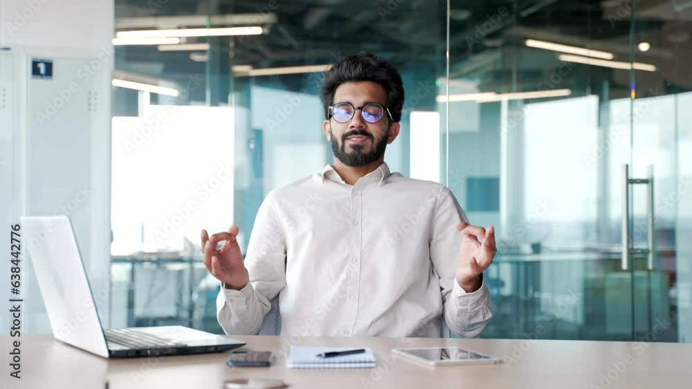 A young bearded businessman meditating with closed eyes during a break while sitting at the workplace in a office. A calm handsome worker rests and feels peace of mind. Male relaxes to relieve stress