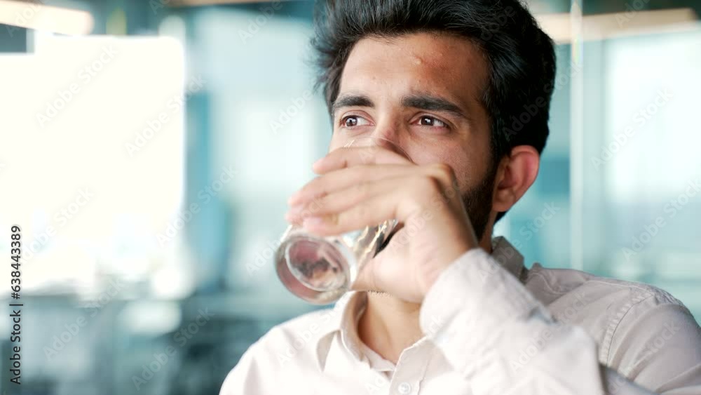 Close up A young bearded man in a shirt drinks water from a glass while sitting at a workplace in the office. Happy smiling employee feeling relieved, enjoying a clean cool drink, relaxing and resting