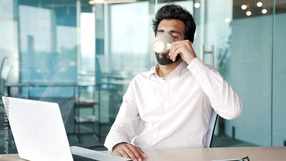 Young man in a shirt drinks tea from a cup while sitting at a workplace in a modern office. Happy businessman enjoys a hot health drink. Smiling employee feeling relief, resting during a break at work Young man in a shirt drinks tea from a cup while sitting at a workplace in a modern office. Happy businessman enjoys a hot health drink. Smiling employee feeling relief, resting during a break at work