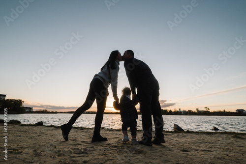 Mother kisses father and daughter near lake in nature. Silhouette at sunset. Spending time together on beach on vacation. Mom, dad hugs child and walk. Concept of autumn holiday. Family photo outdoors