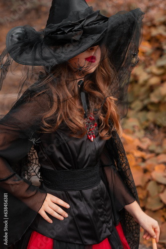 a little girl witch in black and red dress and pointed hat with a veil and spider, standing in yellow leaves, halloween costume
