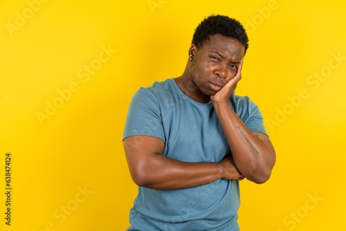 Very bored Young handsome man standing over yellow studio background holding hand on cheek while support it with another crossed hand, looking tired and sick, photo