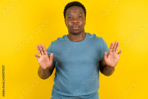 Serious Young handsome man standing over yellow studio background pulls palms towards camera, makes stop gesture, asks to control your emotions and not be nervous photo
