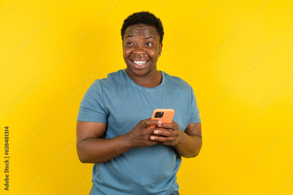 handsome African American standing over yellow studio background holding in hands mobile phone and looking aside with dreamy look. handsome African American standing over yellow studio background holding in hands mobile phone and looking aside with dreamy look.