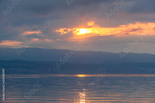 The sun setting behind mountains in Croatia. The Mediterranean Sea is calm and clear, reflecting the sunbeams. The sky is orange and covered with clouds. Many cities along the coastal line. Calmness photo