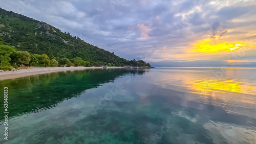 A stony beach along the shore of Medveja in Croatia. The Mediterranean Sea is calm and clear. There is a lush forest with a small town at the shore. The sky is painted yellow. Sunset time. Calmness photo