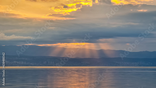 The sun setting behind mountains in Croatia. The Mediterranean Sea is calm and clear, reflecting the sunbeams. The sky is orange and covered with clouds. Many cities along the coastal line. Calmness