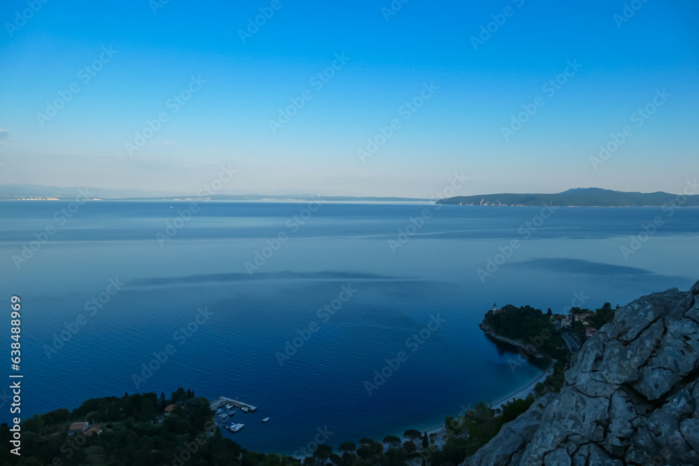 Panoramic view of the shore along Medveja, Croatia seen from above. The town is surrounded by thick forest. A small pier with a few boats. Endless horizon. An island in the back. Summer holidays