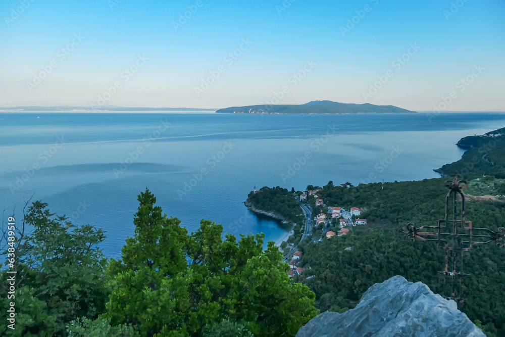 A small metal cross on a rock with a panoramic view of the shore along Medveja, Croatia seen from above. The town is surrounded by thick forest. Endless horizon. An island in the back. Summer holidays