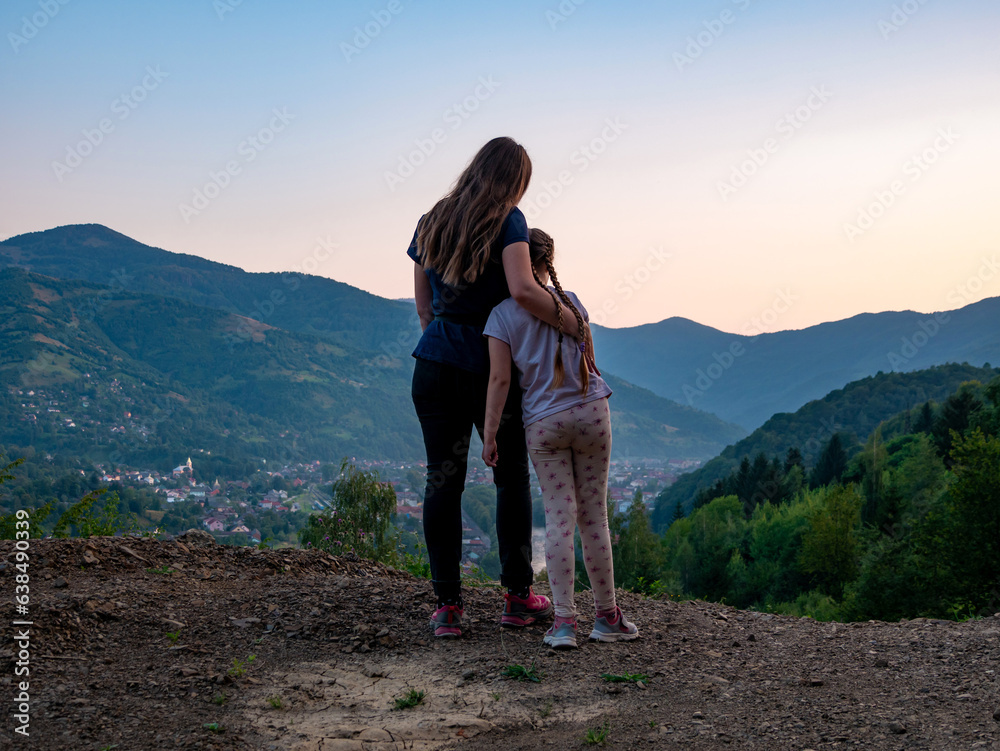 Fototapeta premium Travelers mother and daughter standing back view beautiful Carpathian Mountains background on sunset, having fun spending time together enjoying nature. Active lifestyle family travel tourism concept.
