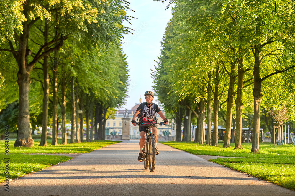attractive senior woman cycling with her electric mountain bike in an old avenue in Ludwigsburg, Baden-Wuerttemberg, Germany with famous Baroque castle in background
