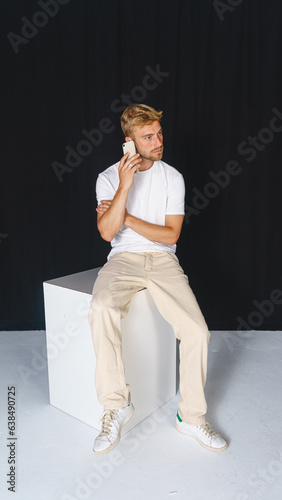 Young, handsome blond man in casual T-shirt and beige pants on black background and sitting talking on the cell phone.