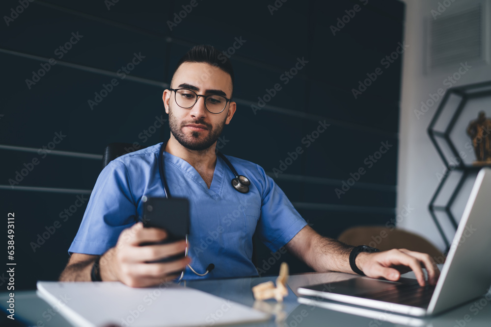 Confident male doctor browsing smartphone while working at laptop Confident male doctor browsing smartphone while working at laptop