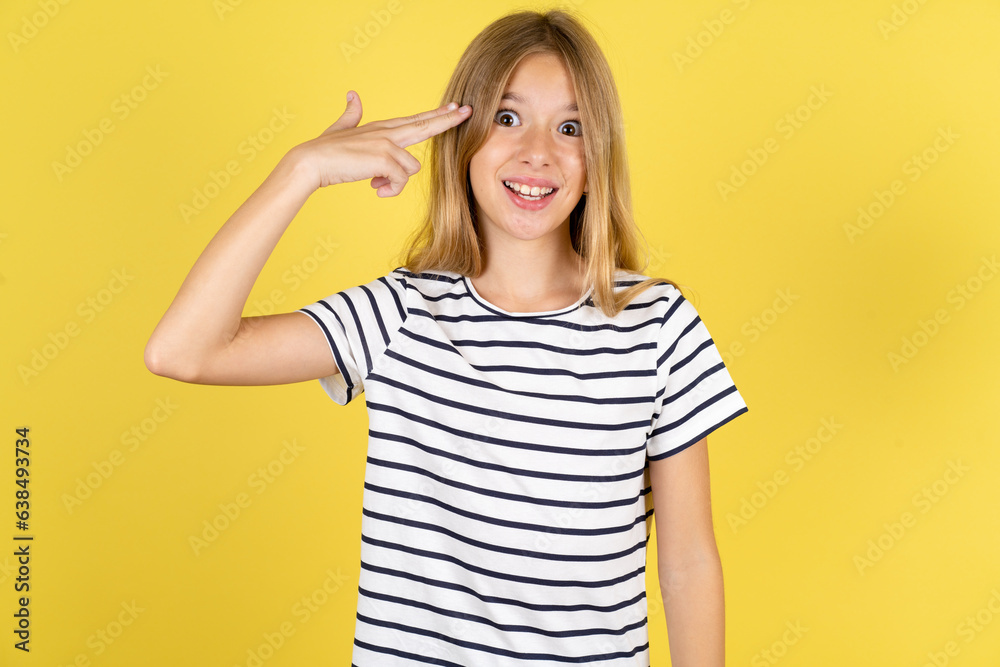 Beautiful caucasian kid girl wearing striped T-shirt foolishness around shoots in temple with fingers makes suicide gesture. Funny model makes finger gun pistol Beautiful caucasian kid girl wearing striped T-shirt foolishness around shoots in temple with fingers makes suicide gesture. Funny model makes finger gun pistol