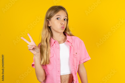 Beautiful caucasian kid girl wearing pink shirt makes peace gesture keeps lips folded shows v sign. Body language concept photo