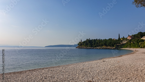 A stony beach along the shore of Medveja in Croatia. The Mediterranean Sea is calm and clear. There is a lush forest with a small town at the shore. Clear, blue sky. Summer remedy. Holidays vibes.