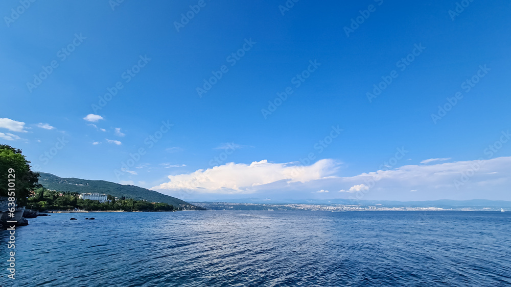 A panoramic view of the shore along Lovran, Croatia. There is a small town located between the lush trees. The Mediterranean Sea has turquoise color. Big boulders in the water. Sunny day. Calmness
