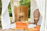 Summer gazebo with flowing white curtains. Wedding boho decoration. Decor outdoor terrace with wicker furniture. Outdoor design of arbour. Rattan peacock armchair and decorative folding screen divider