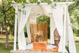 Summer gazebo with flowing white curtains. Wedding boho decoration. Decor outdoor terrace with wicker furniture. Outdoor design of arbour. Rattan peacock armchair and decorative folding screen divider