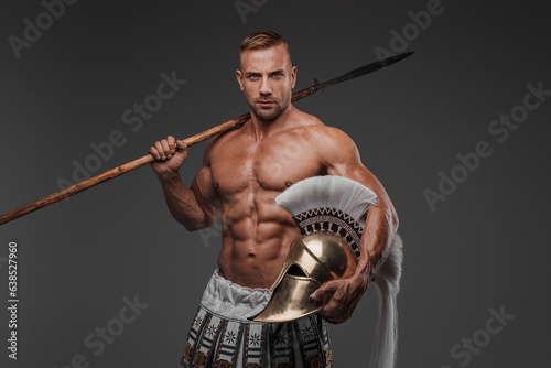 Muscular ancient hoplite warrior with a bare torso and wearing pteruges, posing with a helmet and spear in hand against a grey background