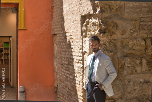 Fototapeta Naklejka Na Ścianę i Meble -  Attractive young businessman with beard, suit and tie, posing with hands in pockets leaning against a stone wall. Concept beauty, fashion, success, achiever, trend.