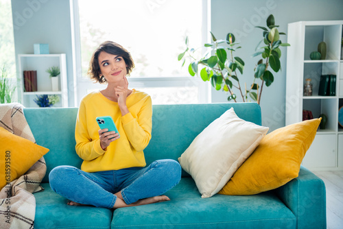 Portrait of pretty dreamy girl sitting on soft cozy couch use phone watching up planning weekend indoor house modern flat