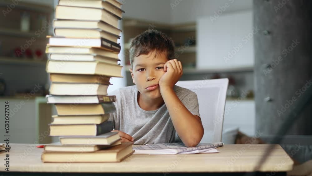 Tired exhausted stressed little boy student looking from behind large pile of books and looking at camera with sad face while preparing for exams at home Overwork education concept Tired exhausted stressed little boy student looking from behind large pile of books and looking at camera with sad face while preparing for exams at home Overwork education concept