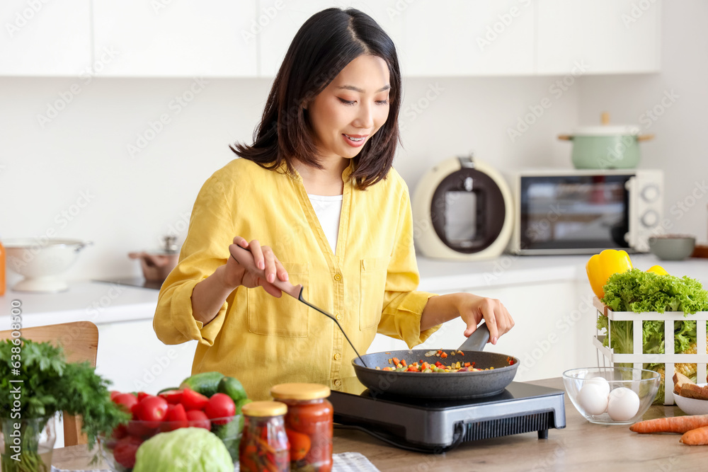 Young Asian woman cooking different vegetables in frying pan on portable electric stove at kitchen