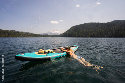 A young woman cools off in an alpine lake at the base of Mt. Hood with her standup paddle board on a summer day. 