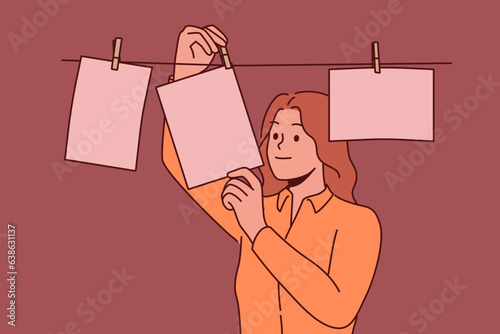 Woman photographer stands in room for developing photographs using old technology and looks at pictures hanging on rope. Girl photographer is fond of creating photos with use of equipment last century