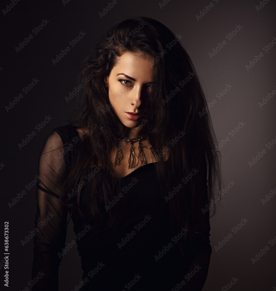 Beautiful woman in black dress posing in fashion necklace on the neck. Long hairstyle on dark shadow background. Toned portrait. Closeup. Beautiful woman in black dress posing in fashion necklace on the neck. Long hairstyle on dark shadow background. Toned portrait. Closeup.