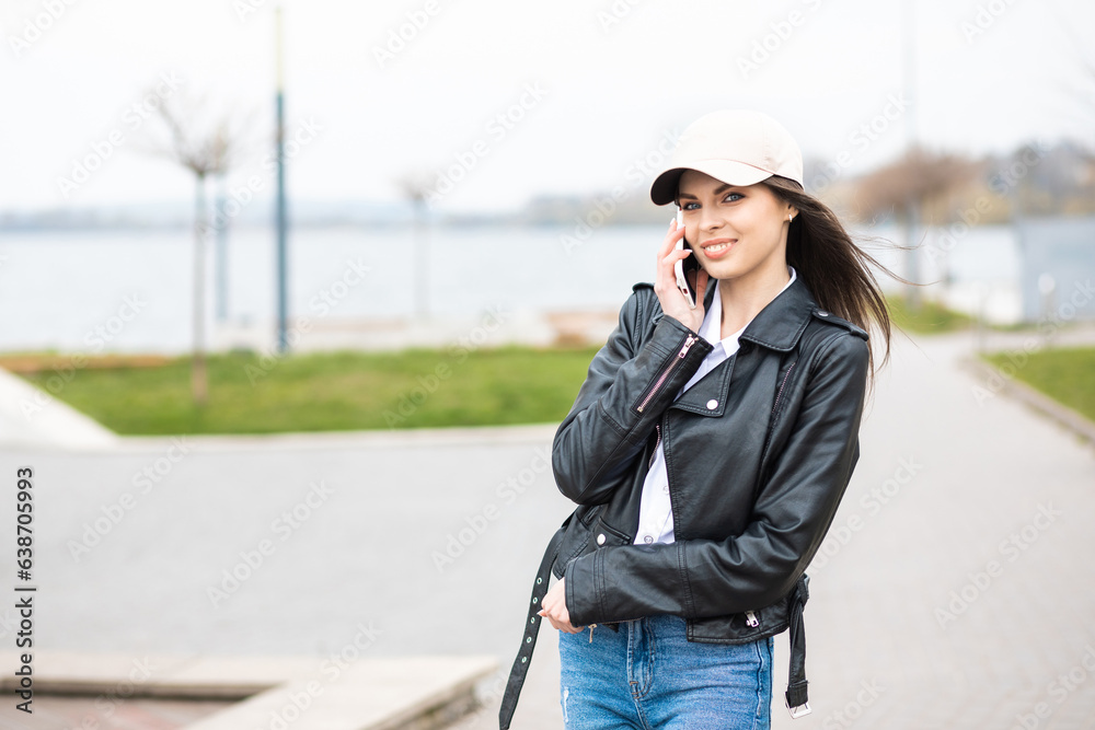 Obraz premium A young pretty girl, dressed in a leather jacket and jeans, spends time alone on the river. The girl holds a mobile phone in her hands and talks