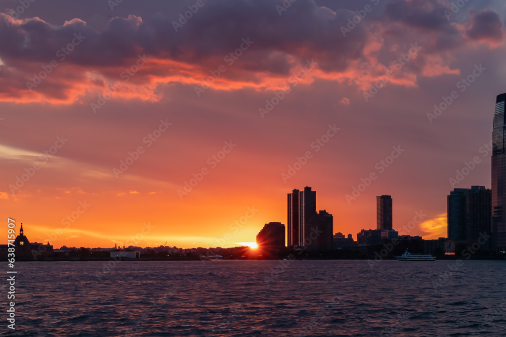 Breath-taking afterglow over New York City skyline and Hudson River at dusk. The sun goes down behind tall skyscrapers. The sky is bursting with orange and yellow. Calmness and relaxation