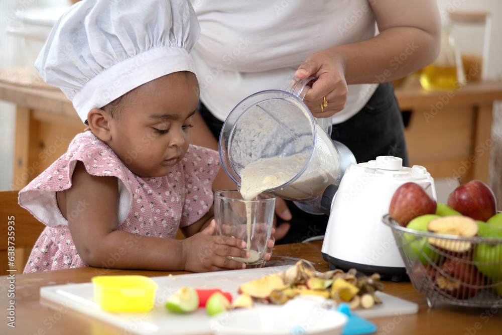 Cute little African girl waiting for her mother's smoothie drink, mom is pouring smoothie into a glass, close up, homemade drink at home Cute little African girl waiting for her mother's smoothie drink, mom is pouring smoothie into a glass, close up, homemade drink at home